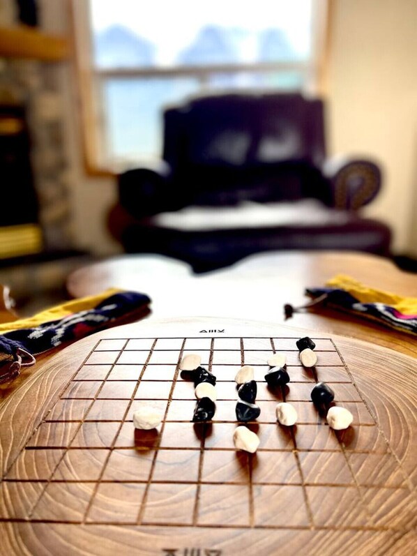 Un tablero de Go con piezas en juego, una ventana con vistas a montañas y un fondo desenfocado con un sofá y un televisor.