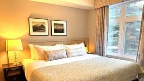 A bedroom with a large bed, a nightstand with a lamp, two framed pictures on the wall, and a window with curtains.