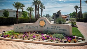 A sign for Cas Palmas Resort Condominiums with a sun emblem, surrounded by a flower bed and palm trees. - 3BR Resort Oasis: Pools, Gym & Sunset Views (St. George)