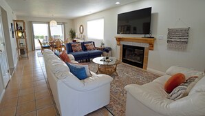 A spacious living room with a fireplace, a flat-screen TV mounted above it, and a large window providing natural light. - 3BR Resort Oasis: Pools, Gym & Sunset Views (St. George)