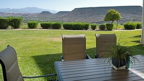 An outdoor seating area with two chairs and a table, a potted plant, and a view of a grassy landscape with hills in the background. - 3BR Resort Oasis: Pools, Gym & Sunset Views (St. George)