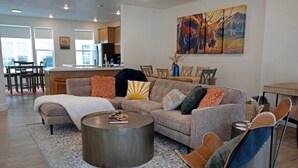 A modern living room with a sofa, a coffee table, and a chair. There is a painting on the wall and a kitchen in the background. - Modern 3BR Retreat | Mtn Views & Garage Parking (South Salt Lake)