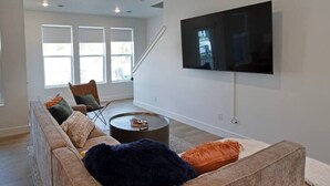 A modern living room with a large flat-screen TV mounted on the wall, a beige sectional sofa with multiple cushions, a coffee table, and a chair. - Modern 3BR Retreat | Mtn Views & Garage Parking (South Salt Lake)