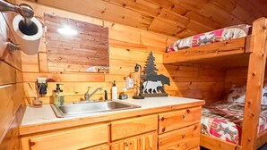 A wooden cabin interior with a kitchen area, a bunk bed, and a handprint rug.