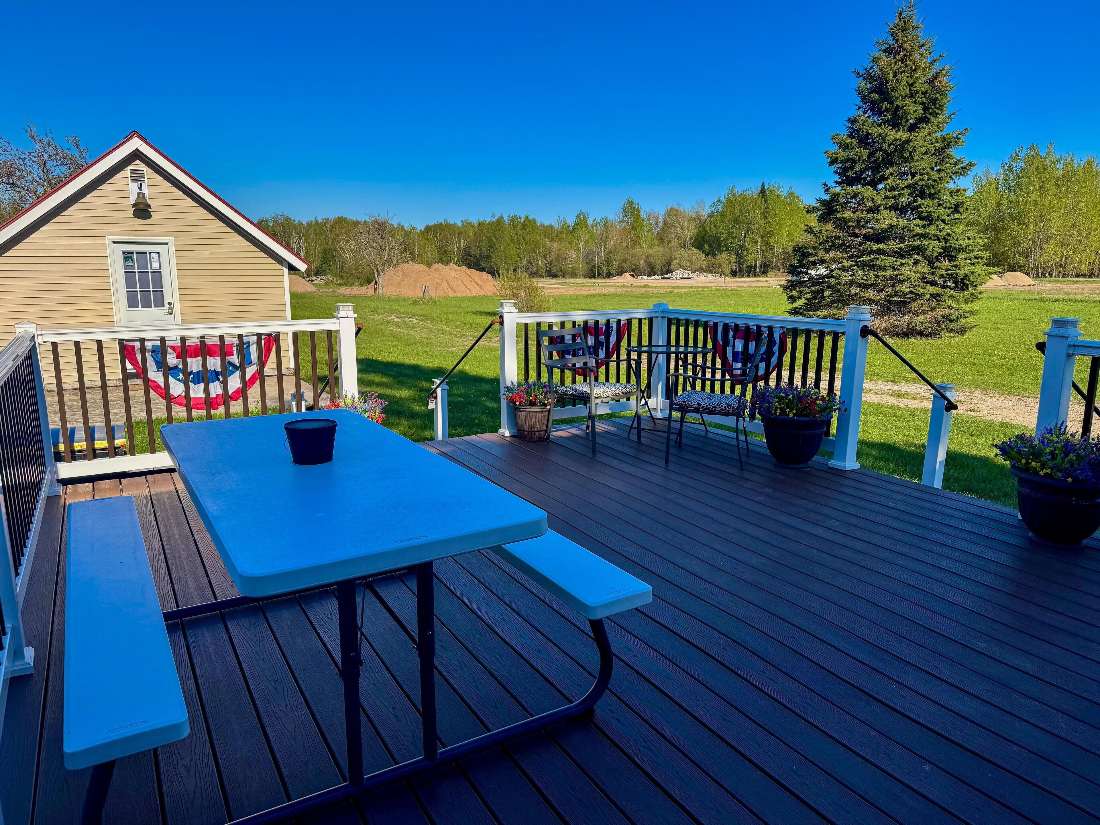 Une terrasse avec une table de pique-nique et des chaises, un drapeau, et une vue sur un champ verdoyant et des arbres.
