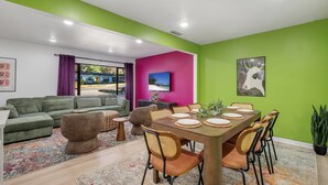 A modern living room with a green wall, a wooden dining table set for four, a green sofa, and a large window. - Spacious 4BD - Central Stay w/ Backyard & Games (Tallahassee)