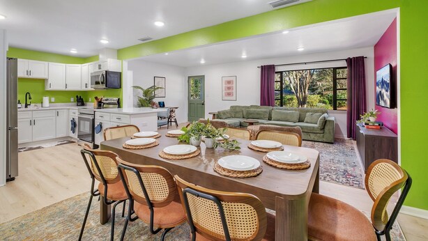 A modern kitchen with white cabinets, a dining area with a wooden table set for six, and a living area with a green sofa and a TV mounted on the wall. - Spacious 4BD - Central Stay w/ Backyard & Games (Tallahassee)
