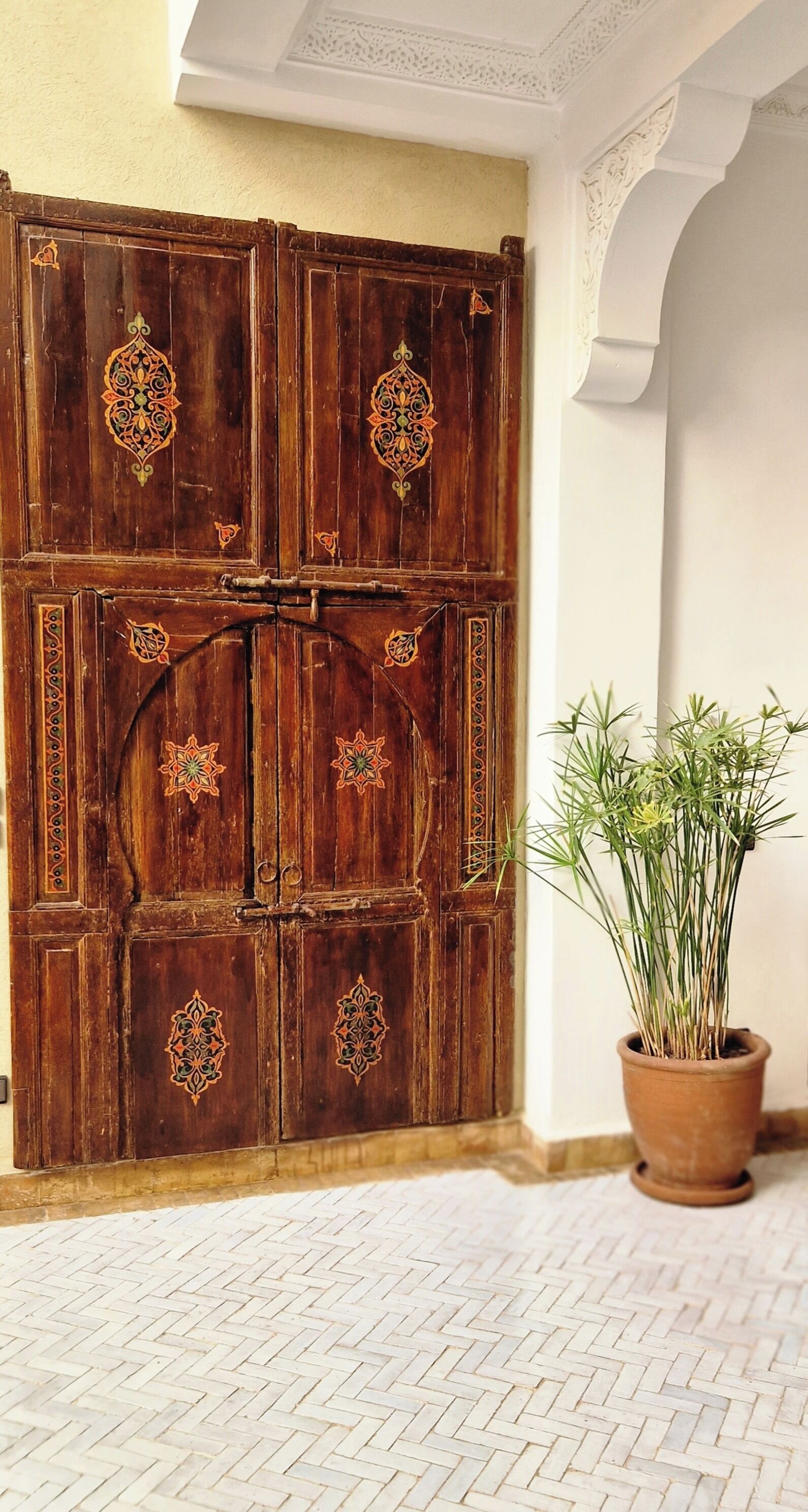 Una puerta de madera con elaborados grabados, una planta en maceta y un suelo con estampado tipo espiga.