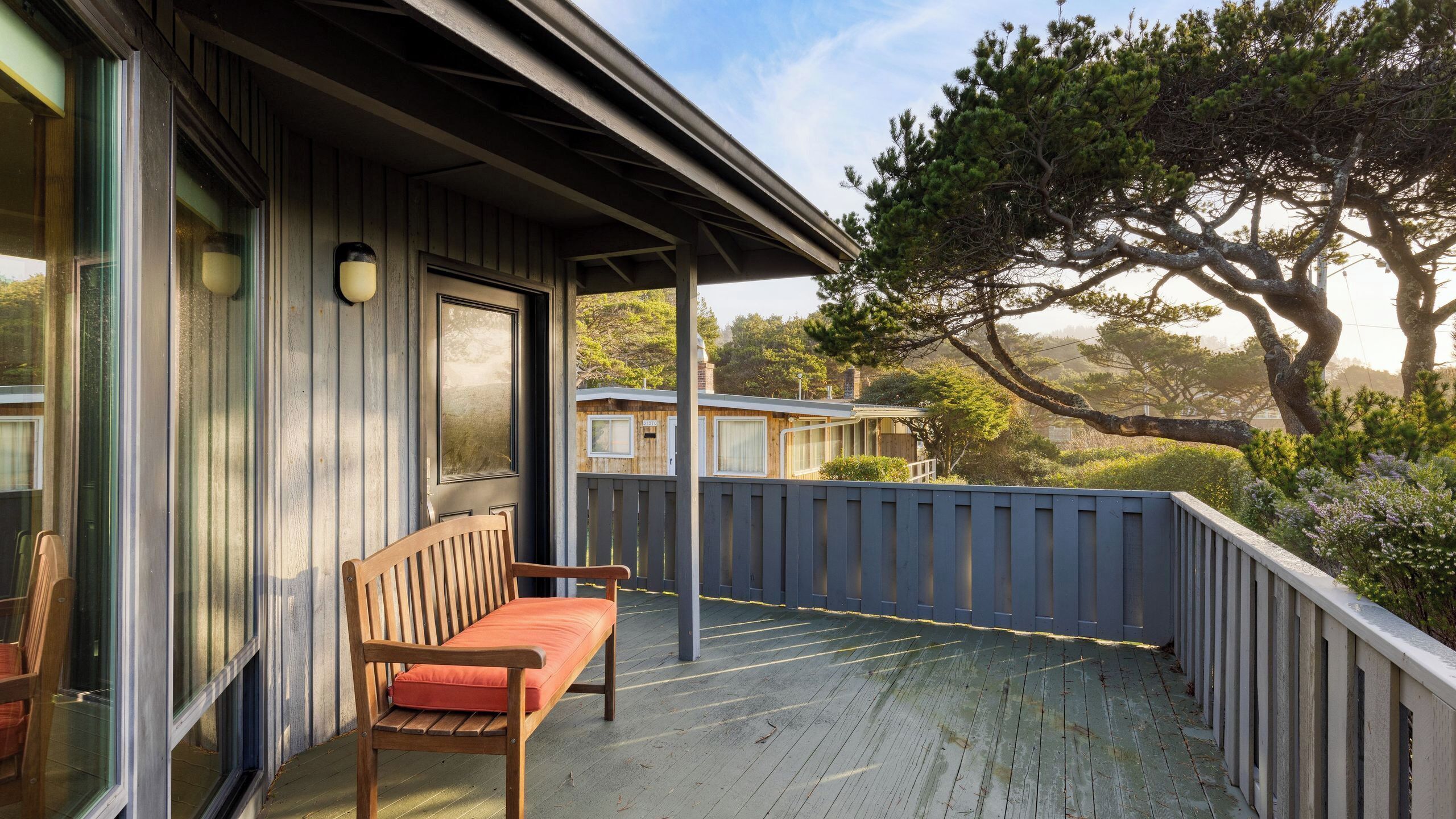 A wooden deck with a bench, a covered area with a roof, and a view of trees and a building in the background.