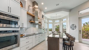A modern kitchen with white cabinetry, stainless steel appliances, and a central island with bar stools. - Luxury Waterfront Retreat - 6BR, Heated Pool, Walk to Beach (Bonita Springs)