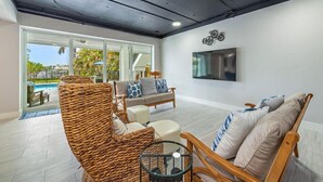 A modern living room with wicker furniture, a flat-screen TV, and a view of a pool. - Luxury Waterfront Retreat - 6BR, Heated Pool, Walk to Beach (Bonita Springs)