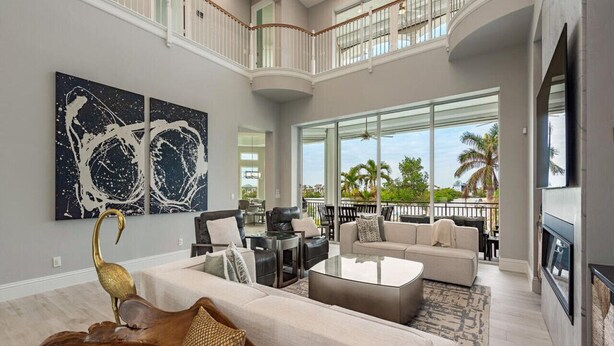 A spacious living room with a high ceiling, a balcony with a view of palm trees, and a large painting on the wall. - Luxury Waterfront Retreat - 6BR, Heated Pool, Walk to Beach (Bonita Springs)