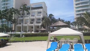 Un hotel frente a la playa con varios pisos, palmeras y zonas de estar al aire libre.