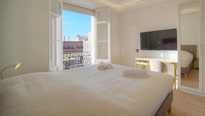 A bedroom with a large bed, a TV, and a balcony with a view of buildings.