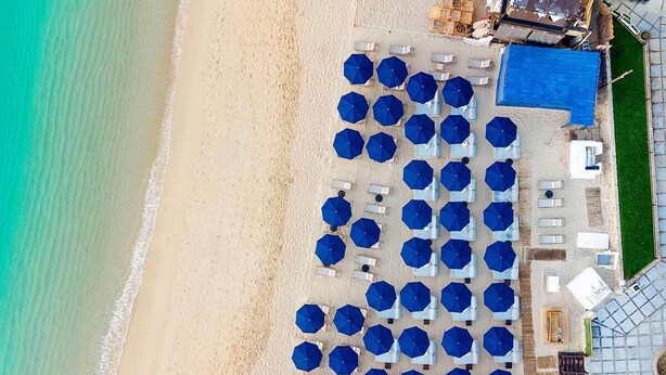An aerial view of a beachside hotel with rows of blue umbrellas and white lounge chairs, a pool area, and a building with a blue roof. - Chic 2-bedroom deluxe suite. A few steps away from Medano Beach!! CABO SAN LUCAS (Cabo San Lucas)