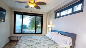 Une chambre à coucher avec un lit, un ventilateur de plafond, une fenêtre donnant sur des palmiers et une table de chevet avec une lampe.