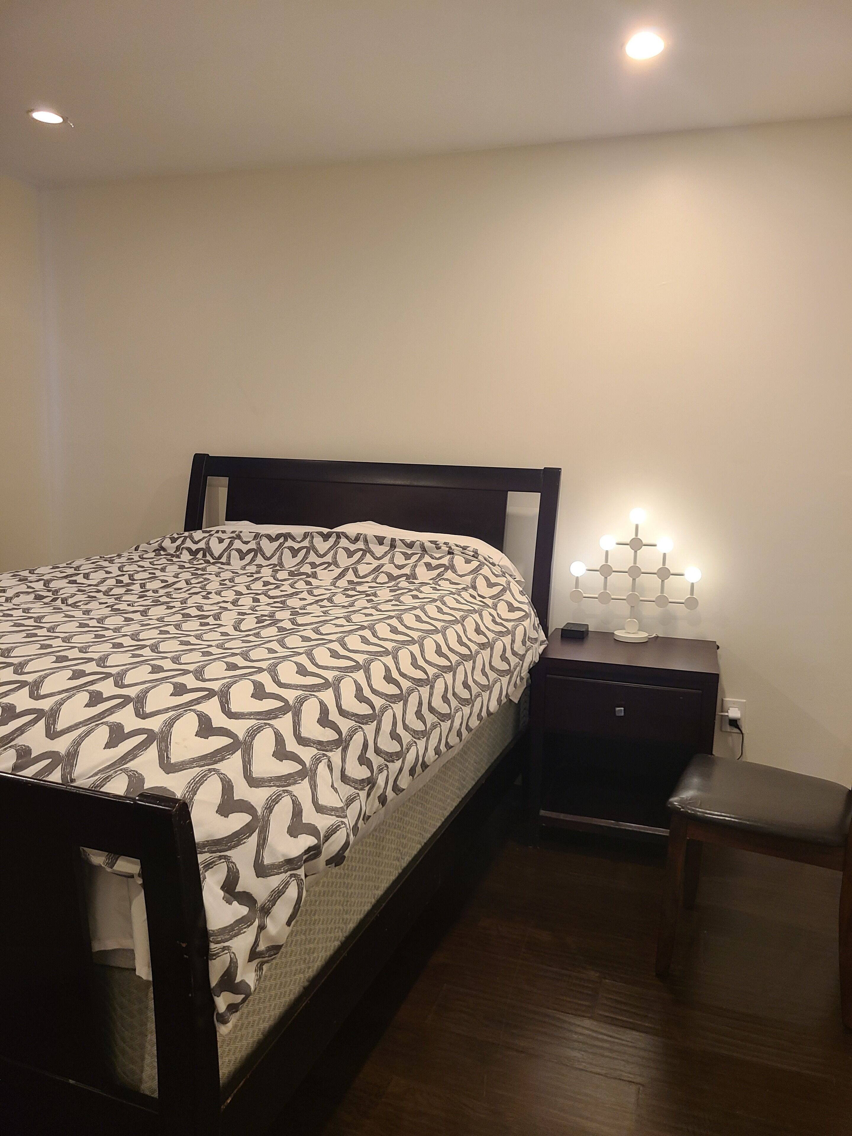 Een net opgemaakt bed met een bedspreider met patroon, een nachtkastje met een lamp en een klein houten krukje.