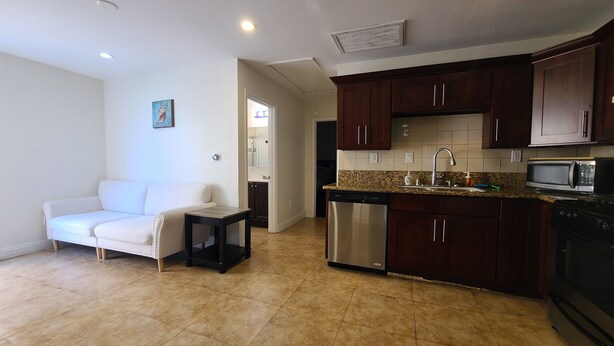 A compact kitchen with dark wooden cabinets, a stainless steel dishwasher, a microwave, and a sink with a granite countertop. - Cozy Cottage, 2-bedroom duplex in delightful Beach City with WiFi, AC (Torrance)