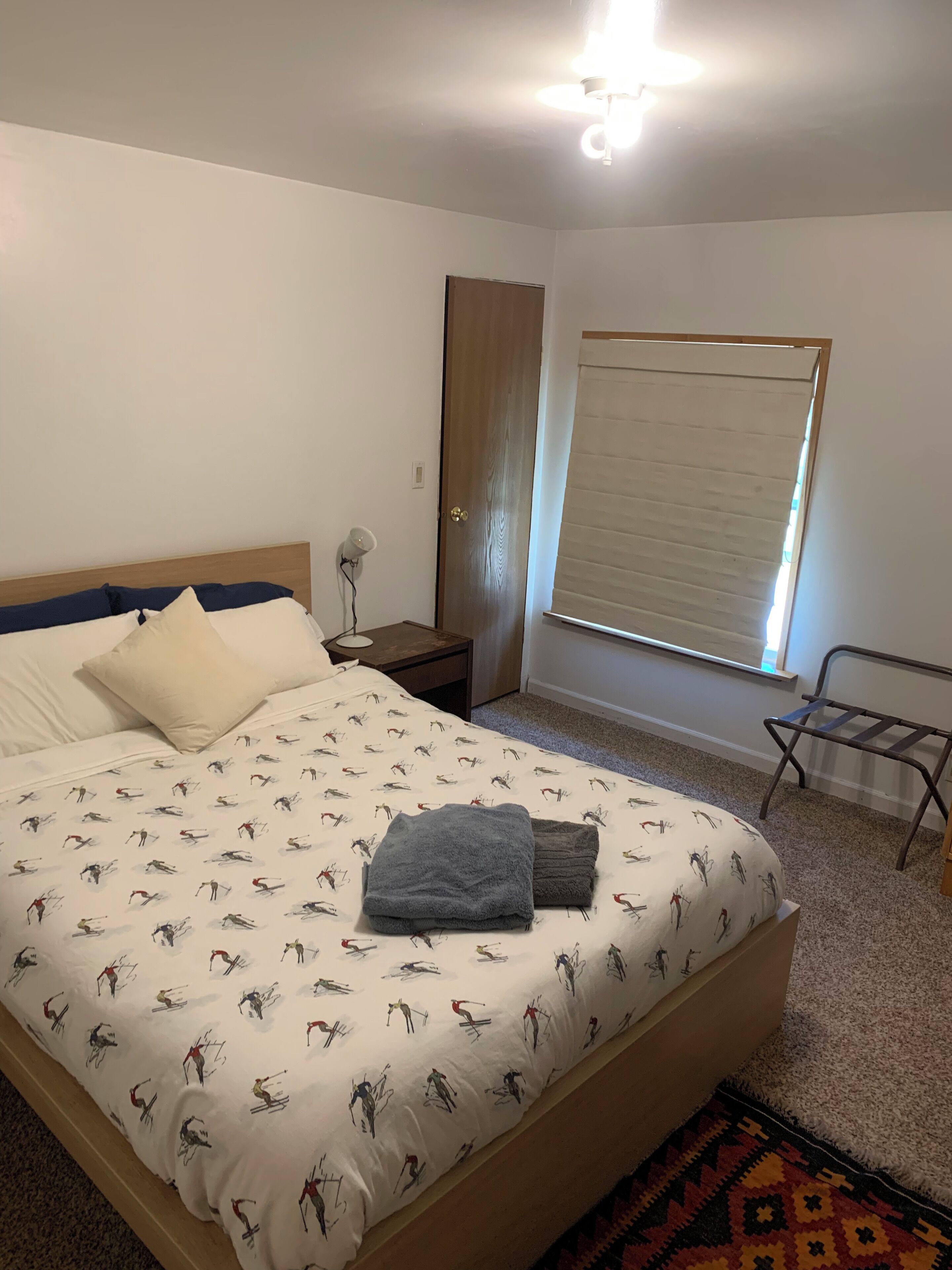 Un dormitorio con cama, mesita de noche, ventana con persianas y una lámpara de pie.