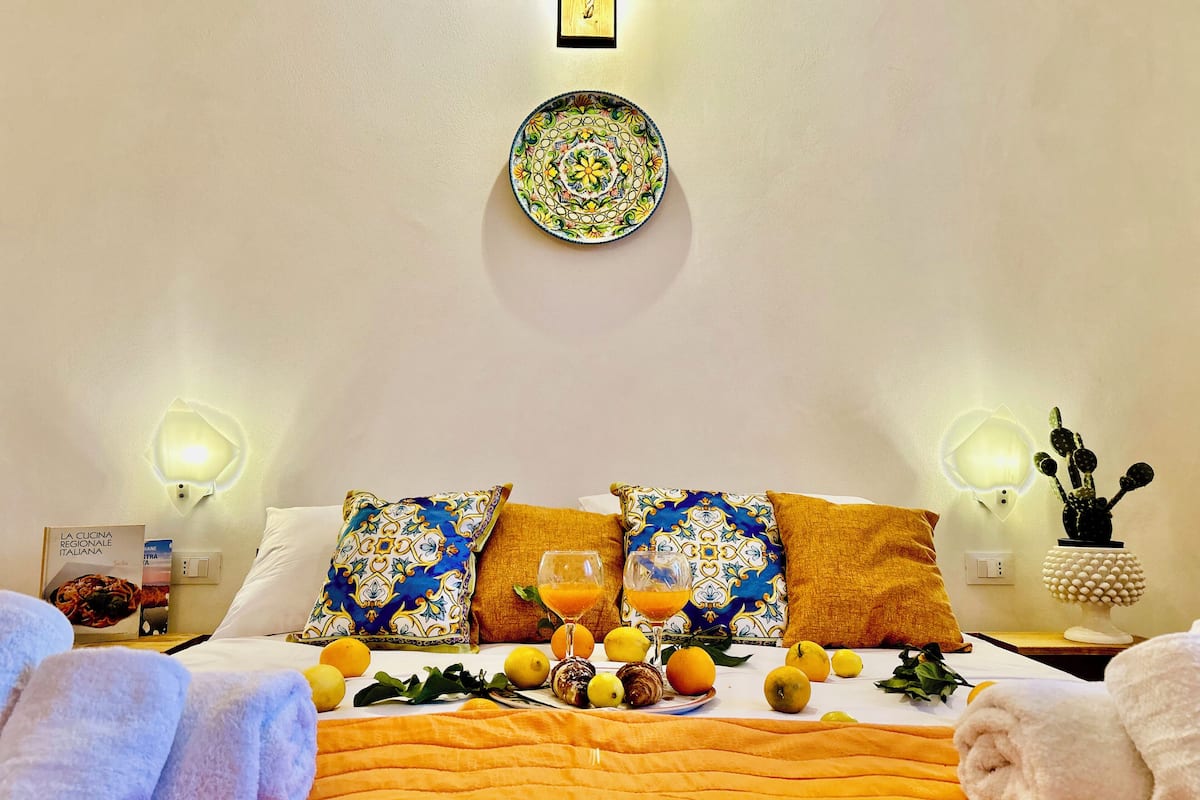 A hotel room with a neatly made bed, a plate with a colorful design on the wall, and a small table with a glass of orange juice and a bottle.