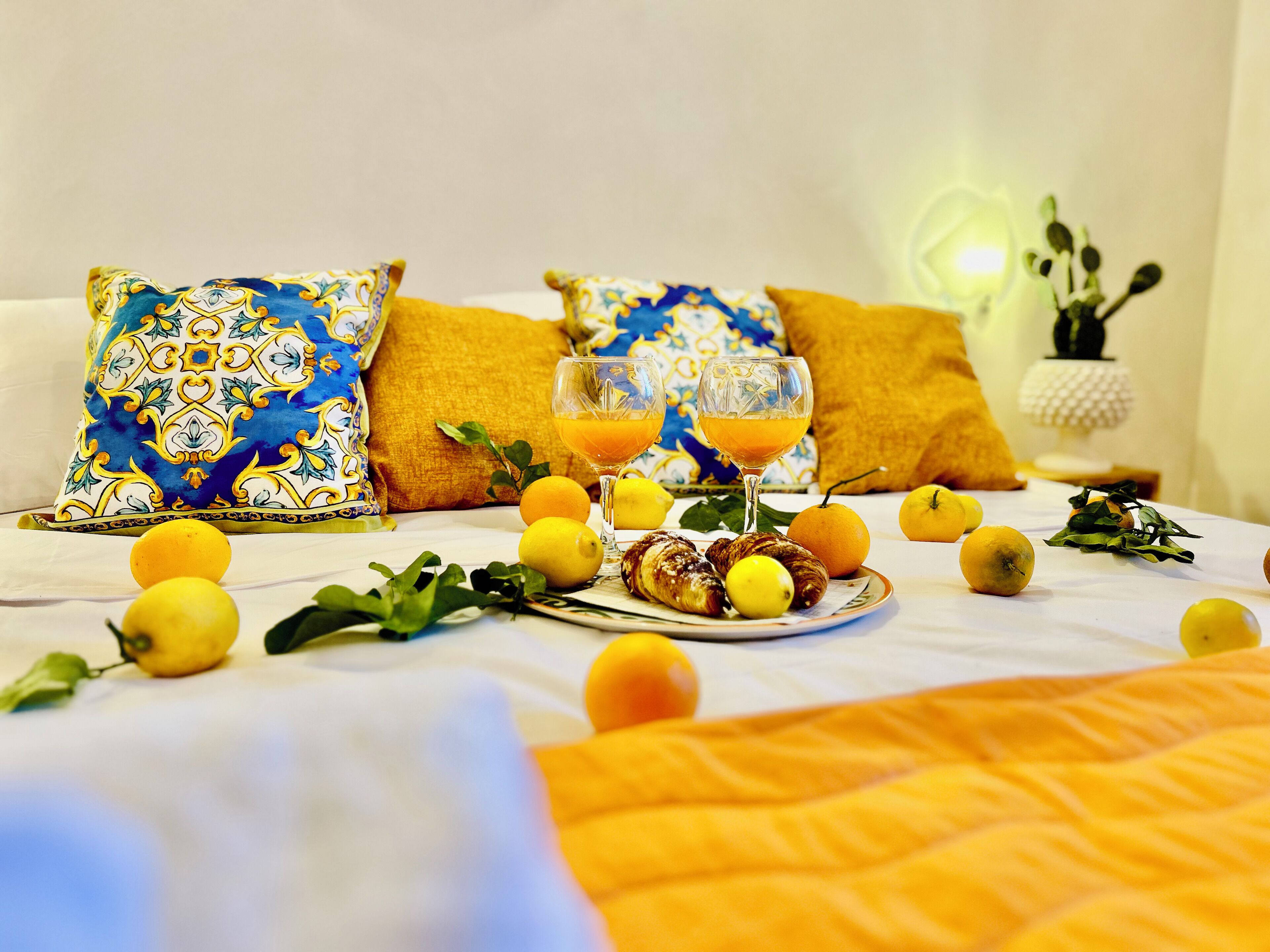Una cama con colcha blanca y amarilla, dos almohadas con diseños azules y amarillos, un plato con cruasanes y limones, y dos vasos de zumo de naranja.