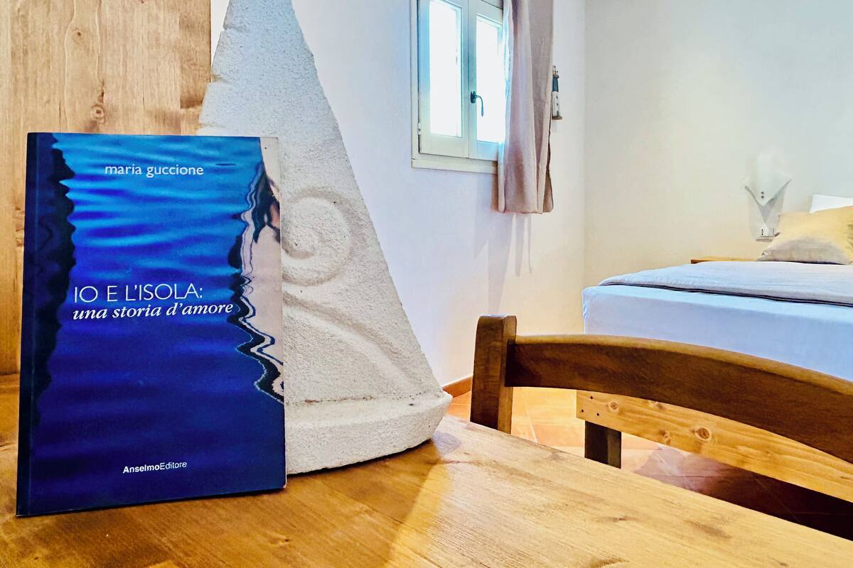 A book titled "IO E L'ISOLA: una storia d'amore" by Maria Guccione on a wooden table in a room with a bed, a window with curtains, and a wall-mounted lamp.