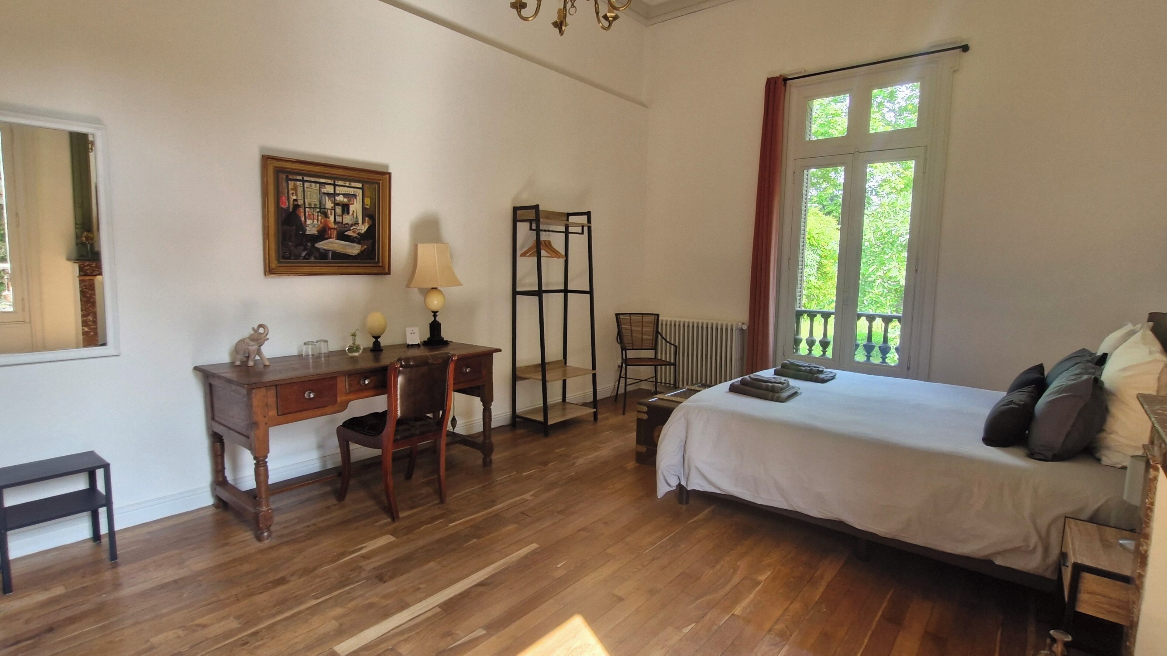 Un dormitorio con un escritorio de madera, una silla, una cama con almohadas, un radiador y una ventana con vista a follaje.