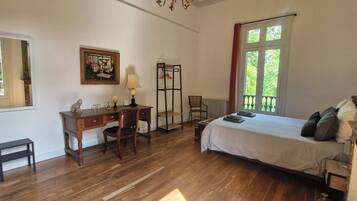 Un dormitorio con un escritorio de madera, una silla, una cama con almohadas, un radiador y una ventana con vista a follaje.