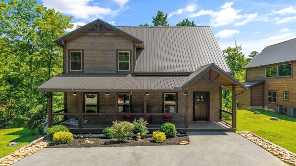 A two-story wooden house with a covered porch, a front yard with landscaping, and a clear sky.