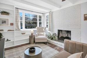 A living room with a white brick fireplace, a white armchair, a sofa, a plaid rug, and a small table with a lamp.