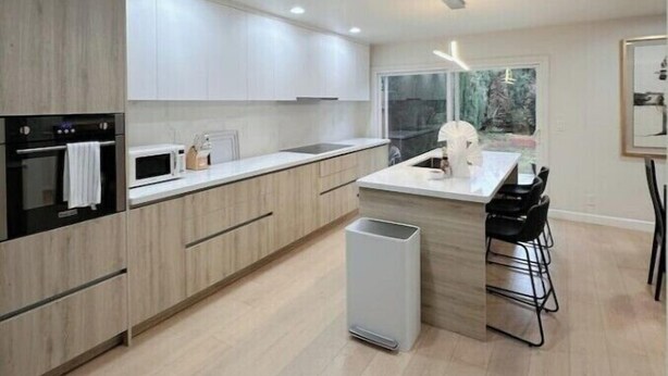 A modern kitchen with light wood cabinets, a central island with bar stools, a built-in oven, and a large window providing a view of the outdoors. - Luxury 3B house near Tec Hubs, High speed WiFi, Chef's Kitchen (San Jose)