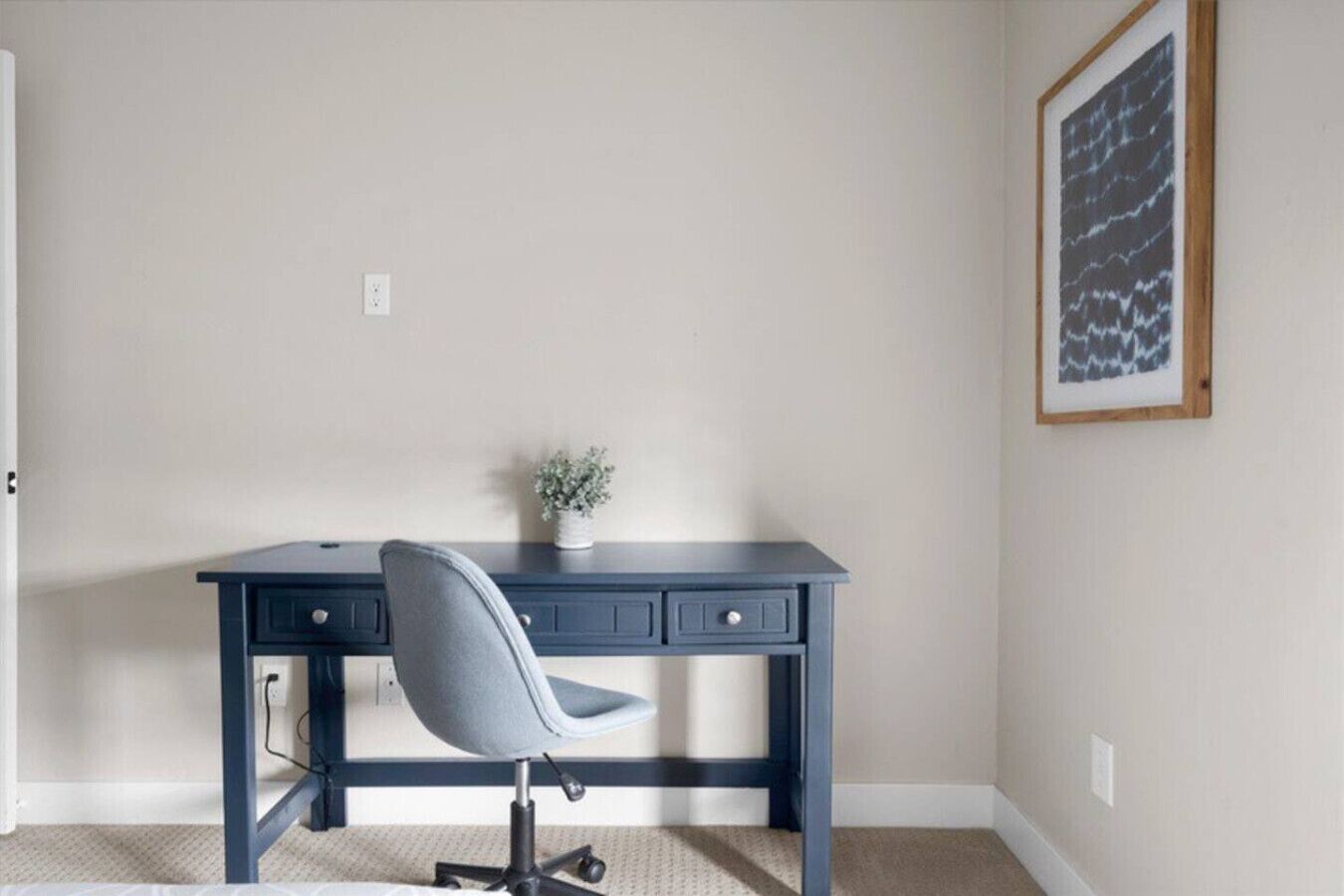 A room with a dark blue desk, a grey office chair, a small potted plant, and a framed abstract artwork on the wall.