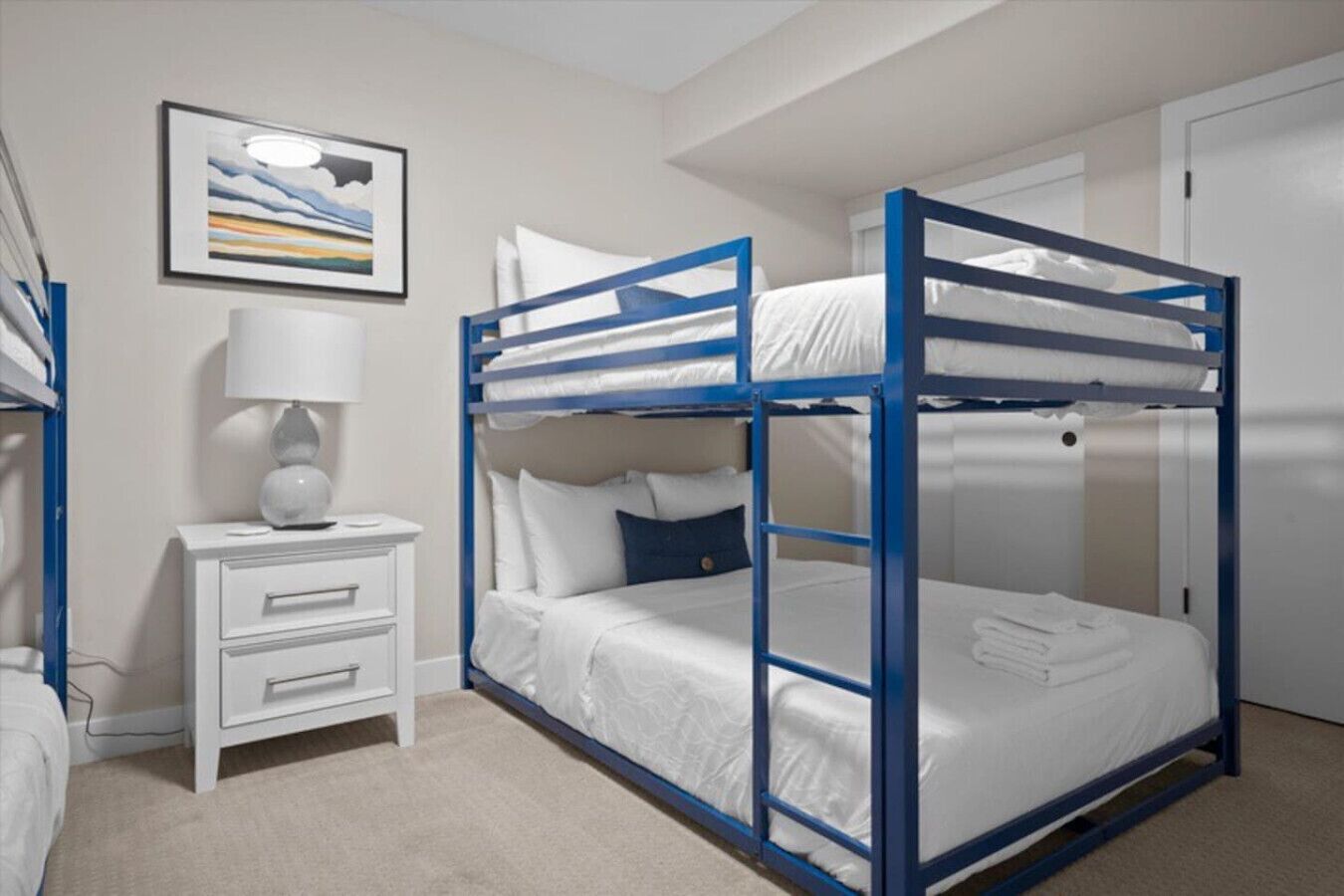 A bunk bed room with a white nightstand, a lamp, and a framed picture on the wall.