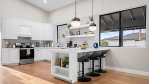 A modern kitchen with white cabinetry, a central island with bar stools, pendant lighting, and a view through a large window. - Pickleball-Basketball-HeatedPool-Spa-2 Primary BRs (Scottsdale)