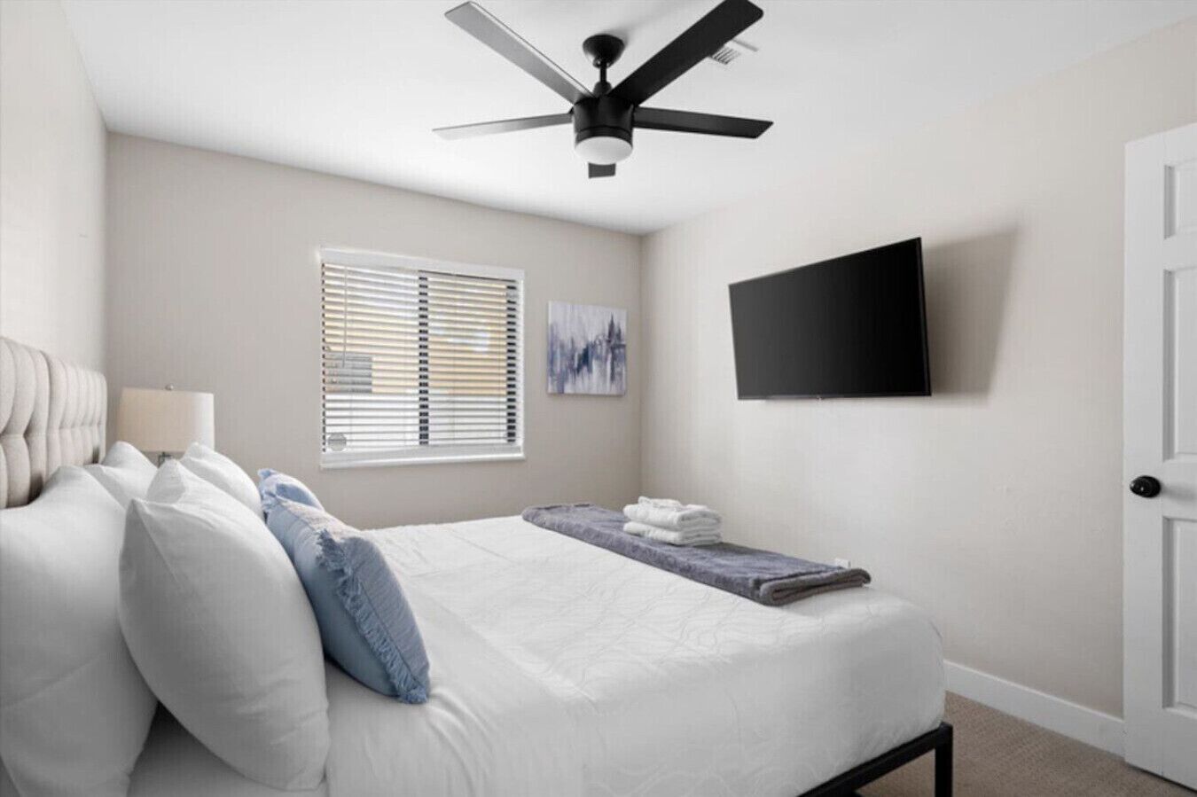 A bedroom with a bed, a ceiling fan, a window with blinds, a wall-mounted TV, and a door.