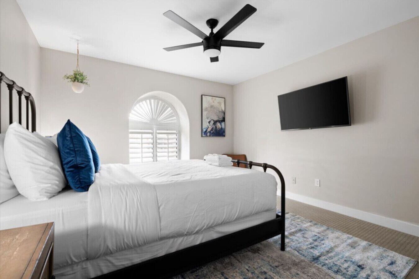 A bedroom with a bed, a ceiling fan, a TV, a window with blinds, and a hanging plant.