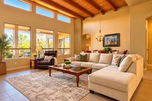 A spacious living room with a sectional sofa, a leather armchair, and a coffee table. There are multiple windows allowing natural light, and a chandelier hangs from the ceiling.