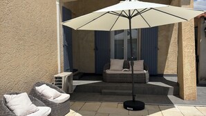 Een overdekt terras met twee rieten stoelen, een witte parasol en een rieten bank met kussens.