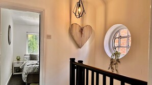A hallway with a staircase, a heart-shaped wall decoration, a round window, and a bed with a view of greenery.