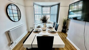 A dining area with a table set for four, a large window with blinds, a radiator, and a TV on the wall.