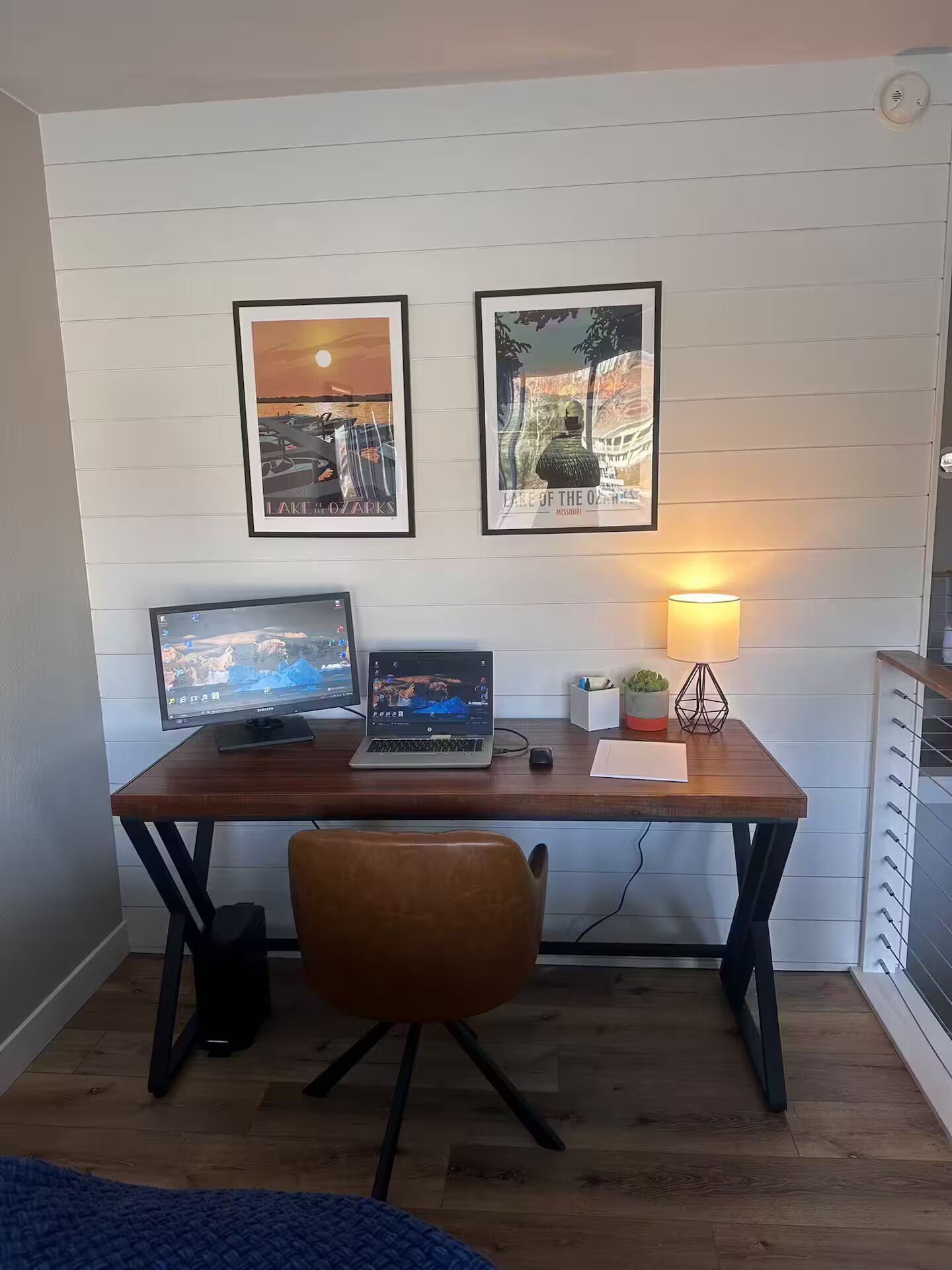 Een werkplek met een houten bureau, twee monitoren, een laptop, een lamp en een lijst met een landschap.