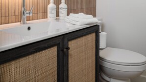 Une salle de bain moderne avec un lavabo blanc, un miroir rond et des toilettes. On peut voir deux flacons de savon pour les mains et le corps posés sur le lavabo.