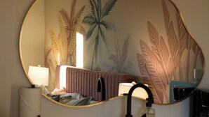 Une chambre avec un lit, un miroir et un mur décoré d’une fresque représentant des feuilles tropicales.