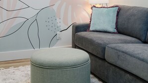 Ett vardagsrum med en grå soffa, en turkosfärgad kudde, en rund pall och en vägg dekorerad med abstrakt blomstermotiv.