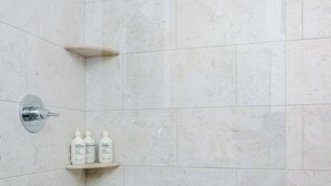 A bathroom with white tiles, a showerhead, a handheld shower, and a shelf with three bottles of shampoo. - Aviles Double Full- 5 min walk to beach! (St. Augustine)