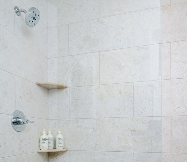 Un baño con azulejos blancos, cabezal de ducha, ducha de mano y un estante con tres botellas de champú.