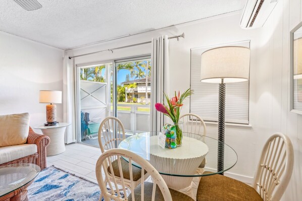 Um quarto claro com mesa de vidro, cadeiras brancas, um sofá de vime e vista para uma área tropical externa.