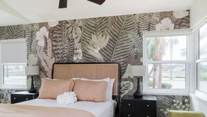 A bedroom with a large bed, two nightstands, a green armchair, and a window with a view of palm trees. - The Ocean Queen Studio -5 min walk to beach! (St. Augustine)