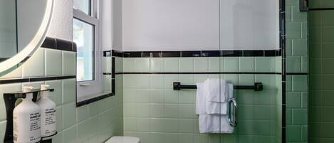 Une salle de bain avec des carreaux verts, des toilettes blanches, un lavabo avec du savon liquide et un miroir.