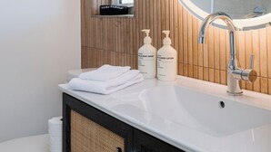 Een badkamer met een witte wastafel, een wand van houten panelen, een spiegel en twee flessen hand- en douchezeep.